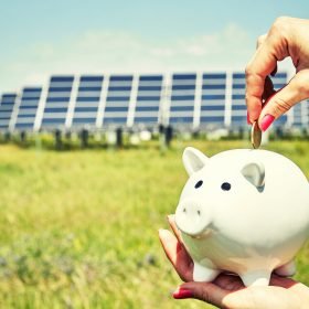 Hand holding money banknote with photovoltaic solar energy panels in background,